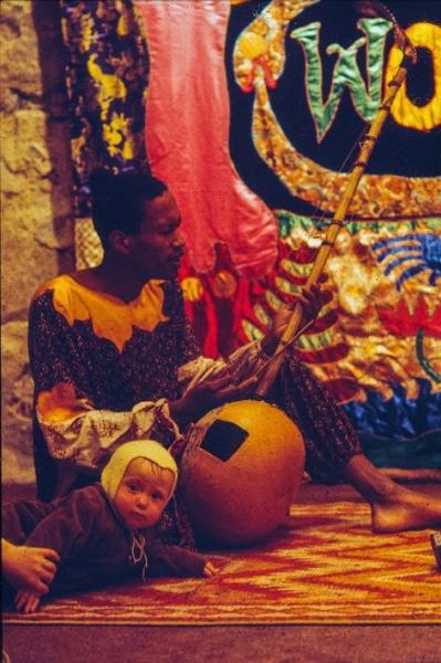 Don Cherry playing donso ngoni while a baby crawls next to him, in front of Eternal Now tapestry by Moki Cherry, at ‘Moki På Galleri 1’, Galleri 1, Stockholm, Sweden, 1973, courtesy of the Cherry Archive, the Estate of Moki Cherry